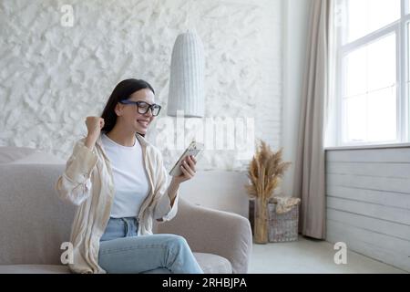 Glückliche junge Frau, die zu Hause auf dem Sofa sitzt und ein Handy benutzt. Er sieht überrascht und fröhlich auf den Bildschirm. Stockfoto