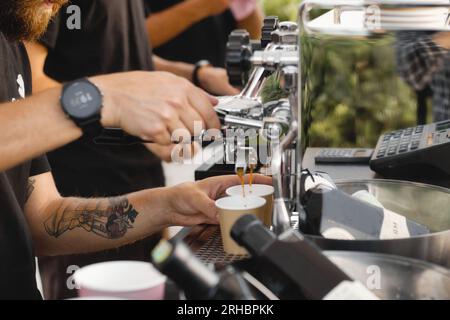 Nahaufnahme der Hände eines tätowierten Barista, der mit einer Kaffeemaschine heiße Getränke in Papierbecher gießt Stockfoto