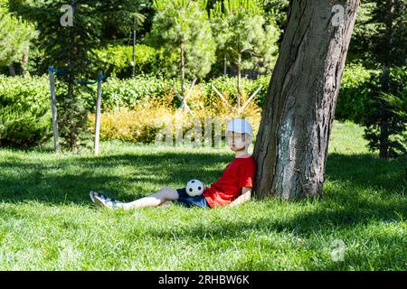 Ein Junge, der im Sommer mit einem Football unter einem Baum sitzt, Georgia Stockfoto