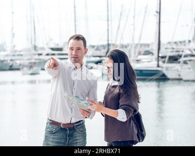 Ein seriöser Mann, der den weiblichen Touristen, der auf der Karte steht, den Weg zeigt, während er auf dem Kai mit festgefahrenen Yachten während des Urlaubs steht Stockfoto