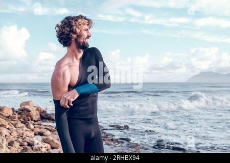 Selbstbewusster junger bärtiger spanischer Sportler mit lockigem Haar in schwarzem Neoprenanzug, der wegschaut, während er sich auf das Surfen im welligen, schaumigen Ozean vorbereitet Stockfoto