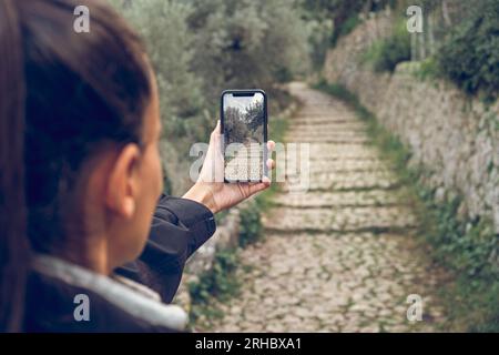 Eine anonyme Wanderer mit Pferdeschwanz im Jackett, die während der Wanderung in Biniaraix auf dem Handy einen engen Steinweg fotografiert Stockfoto