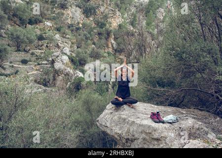 Der ganze Körper einer entspannten jungen hispanischen Wanderin führt eine Padmasana-Pose mit Gebetshändern und gekreuzten Beinen auf einem Felsbrocken auf und übt dabei Yoga Stockfoto