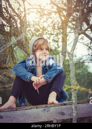 Positiver Barfußjunge in Jeansjacke mit lächelnder Kapuze, die wegschaut, während er auf dem Holzweg inmitten blattloser Baumzweige im Wald sitzt Stockfoto
