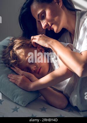 Sanfte Mutter, die das Haar des schlafenden Sohnes hinter dem Ohr versteckt, während sie auf einem bequemen Bett im Schlafzimmer mit hellem Sonnenlicht zu Hause liegt Stockfoto