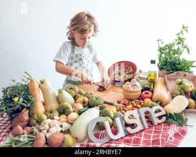 Fokussierter niedlicher Junge in der Schürze schneidet frisches reifes Gemüse auf dem Schneidebrett und bereitet gesunde Lebensmittel aus rohen Zutaten zu Stockfoto