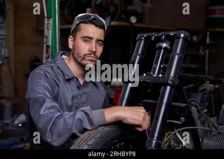 Seriöser, bärtiger, männlicher Mechaniker in Uniform, der in der Nähe des Motorrads sitzt, mit der Hand am Rad sitzt und in die Kamera schaut, während er in einer professionellen Werkstatt arbeitet Stockfoto