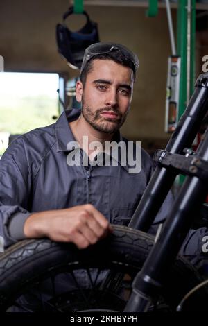 Selbstbewusster Mann in Uniform, der in der Nähe des Motorrads sitzt, mit der Hand am Rad sitzt und in die Kamera schaut, während er in einer professionellen Werkstatt arbeitet Stockfoto