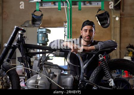 Selbstbewusster bärtiger Techniker trägt Uniform, lehnt sich in der Werkstatt auf das Motorrad und schaut in die Kamera Stockfoto