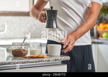 Ein anonymer Mann, der frischen Kaffee aus der Geysirkaffeemaschine in einen Keramikbecher gießt, der auf der Theke neben einem Glas Milch und einem Becher mit braunem Zucker steht Stockfoto