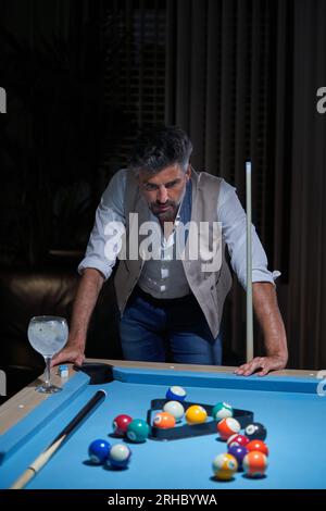 Ein erwachsener Mann, der sich auf dem Billardtisch neben einem Glas Gin Tonic lehnt und in einem dunklen Raum über Bälle nachdenkt Stockfoto