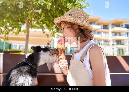 Seitenansicht eines Kindes in legerer Kleidung, das leckere Eiscreme in der Schüssel mit einem Miniatur-Schnauzer-Hund teilt, während es auf einer Holzbank im Stadtpark in der Sonne sitzt Stockfoto