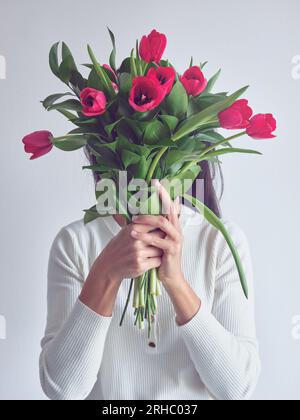Porträt einer unbekannten brünetten Frau im weißen Pullover, die einen Haufen roter Tulpen in den Händen hält, die ihr Gesicht bedecken Stockfoto