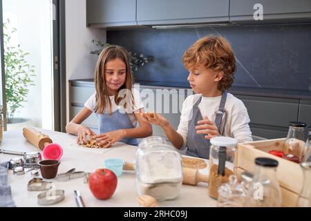 Süße Kinder in Schürzen, die Teig für Kekse zubereiten, während sie am Tisch mit Kochutensilien in der hellen Küche der Wohnung stehen Stockfoto