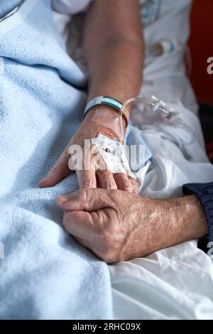 Hochwinkel des anonymen älteren Mannes, der die Hand einer kranken Frau hält und während der Rehabilitationsbehandlung bei Huren einen angeschlossenen Venenkatheter auf dem Bett auflegt Stockfoto