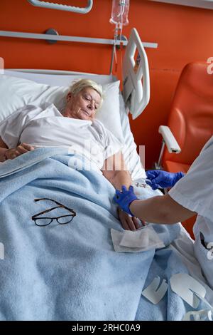 Hochwinkel einer älteren Frau, die auf dem Krankenhausbett liegt und sich Gedanken macht, während eine gesichtslose anonyme Krankenschwester intravenös injiziert Stockfoto