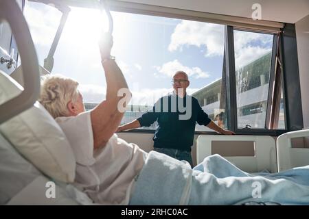 Seitenansicht einer bettlägerigen Patientin mit hängendem Griff im Gespräch mit dem Ehemann während der Behandlung im modernen Leichtkrankenhaus Stockfoto