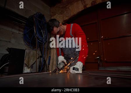 Fokussierter junger bärtiger Arbeiter in rotem Pullover und Jeansanzug, der beim Arbeiten in der Garage ein Stück Eisen schneidet Stockfoto