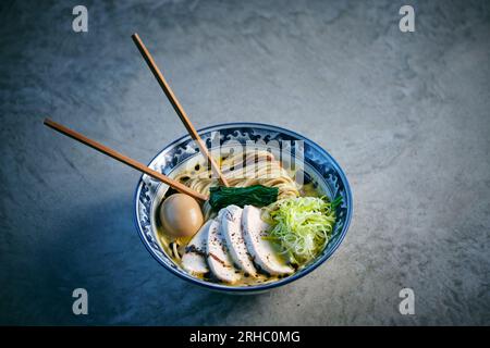 Von oben leckere asiatische Suppe mit gekochten Nudeln und Geflügelstücken mit frischem Gemüse und Essstäbchen auf grauem Hintergrund Stockfoto