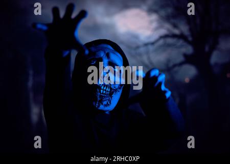 Gruseliges Monster, das die Kamera mit krallenartigen Händen ausstreckend, während es nachts unter blauem Neonlicht durch den dunklen Friedhof schleicht Stockfoto