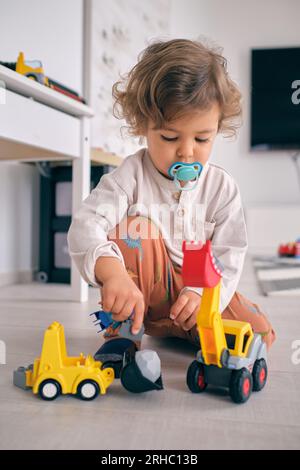 Neugieriger kleiner Junge mit blauem Schnuller, der mit einem Plastiklader spielt, während er zu Hause auf dem Boden im Wohnzimmer sitzt Stockfoto