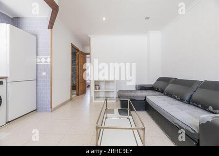 Loft-Apartment mit altem, abgenutztem schwarzen Sofa, Couchtisch und Spiegel aus Metall, weißen Wänden und offener Küche mit weißen Geräten Stockfoto