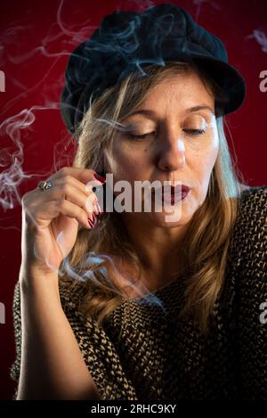 Attraktive und selbstbewusste Frau mit Schwarzmütze. Räucherrauch überall. Studioporträt auf rotem Hintergrund. Stockfoto