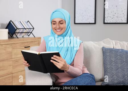 Muslimische Frau liest Buch auf Couch im Zimmer Stockfoto