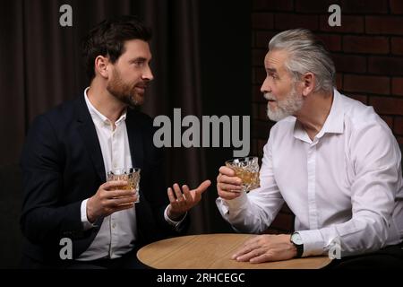 Männer mit Whiskey-Gläsern, die sich drinnen an einem Holztisch unterhalten Stockfoto