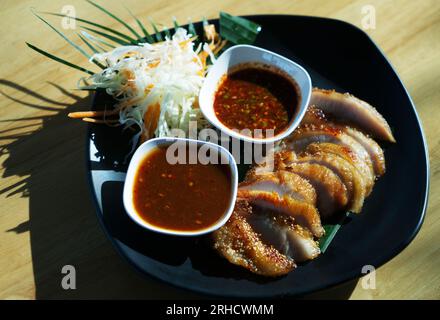 Thailändisches Essen gegrilltes Schweinehals oder Kor Moo Yang in thailändischer Sprache, geschnittenes Schweinefleisch mit zwei Arten Sauce und frischem Salat auf schwarzer Essensplatte auf Holzteller Stockfoto