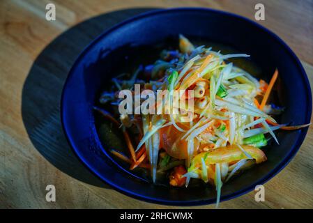 Thailändisches Essen namens Som Tam oder scharfer und saurer frischer Papayasalat, Essen auf schwarzer Essteller auf Holztisch, natürliches Sonnenlicht. Stockfoto