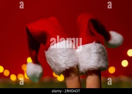 Finger in rot-weißen Weihnachtsmann-Hüten. Zwei glückliche Finger, die das soziale Netzwerk auf rotem Hintergrund repräsentieren. Finger Gesichter in Weihnachtsmannmützen vor rotem Hintergrund. Frohes Familienfest an Weihnachten oder Neujahr. Fingerpaar Stockfoto