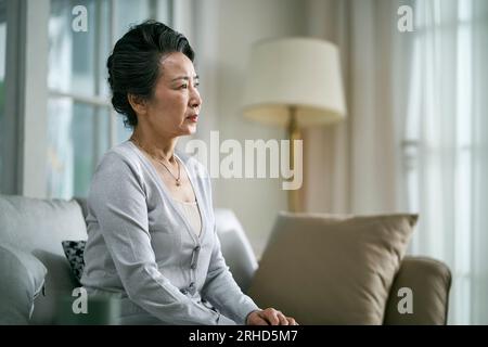 Seitenansicht einer traurigen älteren asiatischen Frau, die allein auf der Couch im Wohnzimmer zu Hause sitzt Stockfoto