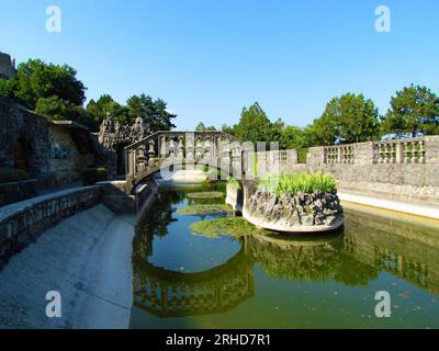 Ovaler Pool im Ferrari-Garten in Stanjel in der Littoral-Region von Slowenien mit einer Steinbrücke und einer kleinen Insel in der Mitte und einem Spiegelbild des bri Stockfoto