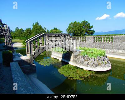 Ovaler Pool im Ferrari-Garten in Stanjel in der Littoral-Region von Slowenien mit einer Steinbrücke und einer kleinen Insel in der Mitte und einem Spiegelbild des bri Stockfoto