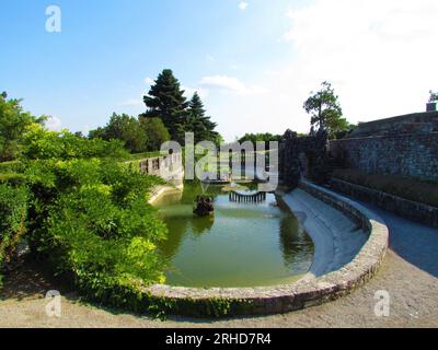 Ovaler Pool im Ferrari-Garten in Stanjel in der Littoral-Region von Slowenien mit einer Steinbrücke und einer kleinen Insel in der Mitte und einem Spiegelbild des bri Stockfoto