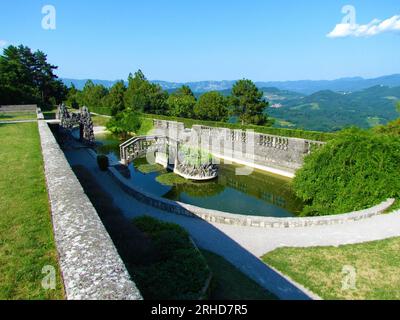 Ovaler Pool im Ferrari-Garten in Stanjel in der Littoral-Region von Slowenien mit einer Steinbrücke und einer kleinen Insel in der Mitte und einem Spiegelbild des bri Stockfoto