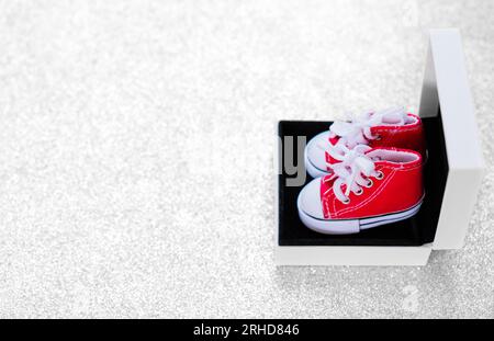 Ein Paar kleine rote Leinwandschuhe in einer kleinen Geschenkbox vor einem glitzernden silbernen Hintergrund. Shopping, Geschenke, Feiertage und E-Comme Stockfoto