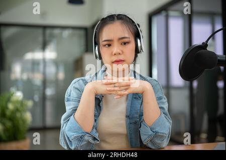 Eine gestresste und ernsthafte asiatische Radiomoderatorin sitzt mit einem unglücklichen Gesicht an ihrem Schreibtisch im Studio, trägt Kopfhörer und hört eine ernsthafte Geschichte f Stockfoto