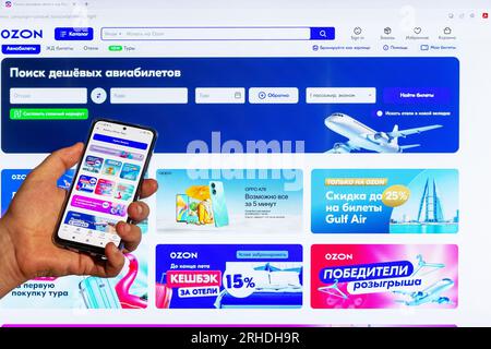 Adygea, Russland - 9. August 2023: Seite des Online-Shops ozon.ru auf einem Computerbildschirm und einem Smartphone in der Hand eines Mannes, der Flugtickets kauft Stockfoto