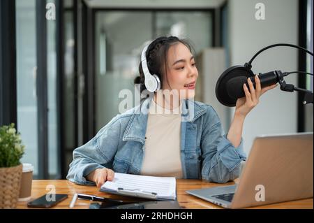 Eine wunderschöne asiatische Radiomoderatorin spricht in ein Mikrofon und gibt während ihrer Sendung interessante Nachrichten an ihr Online-Publikum weiter. Stockfoto