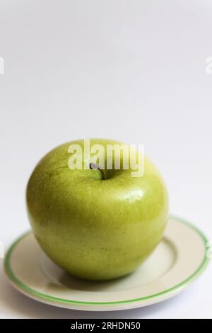 Ein einzelner grüner Apfel, bedeckt mit Wassertropfen auf einer kleinen weißen Platte vor weißem Hintergrund Stockfoto