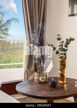 Glasvase mit Eukalyptusblättern, Schmuckkästchen und Räucherstäbchen auf dem Kaffeetisch im Wohnzimmer in der Nähe des Fensters Stockfoto