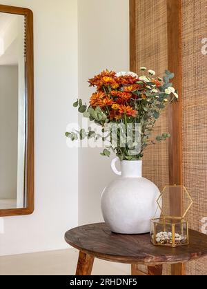 Strauß von orangefarbenen Chrysanthemen mit Eukalyptusblättern auf dem Kaffeetisch im Innenbereich neben dem Spiegel Stockfoto