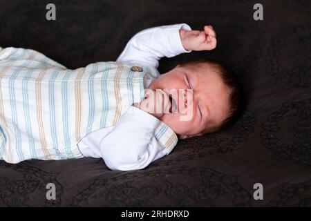 Müdes, verschlafenes Baby, das auf dem Bett der Eltern mit der schwarz gesteppten donna gähnt und sich ausstreckt Stockfoto