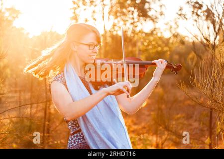 Schöne junge Frau in ihren Teenagern, die ihre Geige draußen zwischen Bäumen spielt und nach ihrer eigenen Melodie tanzt Stockfoto