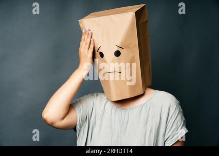 Frau mit der Hand auf dem Kopf und aufgeregtem Lächeln auf dem Papierbeutel auf dem Kopf. Schlechtes Gedächtnis, Emotionskonzept. Stockfoto