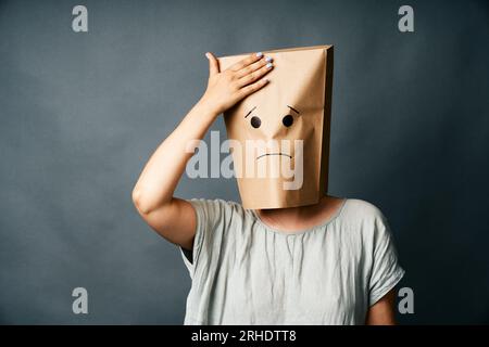 Frau mit der Hand auf dem Kopf und aufgeregtem Lächeln auf dem Papierbeutel auf dem Kopf. Schlechtes Gedächtnis, Emotionskonzept. Stockfoto