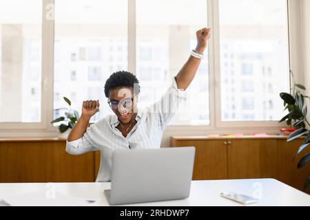 Glückliche und glückliche junge afroamerikanische Büroangestellte, Kollegin, freischaffende Mitarbeiterin, die sich euphorisch fühlte, wenn sie den Laptop-Bildschirm betrachtete, überfreuliche multirassische Frau, die ein Stellenangebot erhielt, machte einen guten Deal Stockfoto