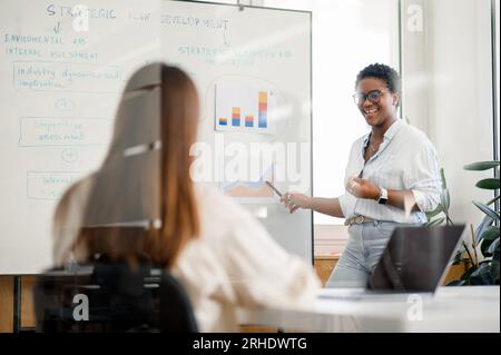 Kluge und qualifizierte afrikanische Geschäftsfrau, Coach, Führungskraft in Brillenfassung, die vor einer Gruppe von Mitarbeitern eine neue Strategie auf dem Whiteboard vorstellt, motivierte Rednerin auf dem Flipchart, die Schulungen durchführt Stockfoto